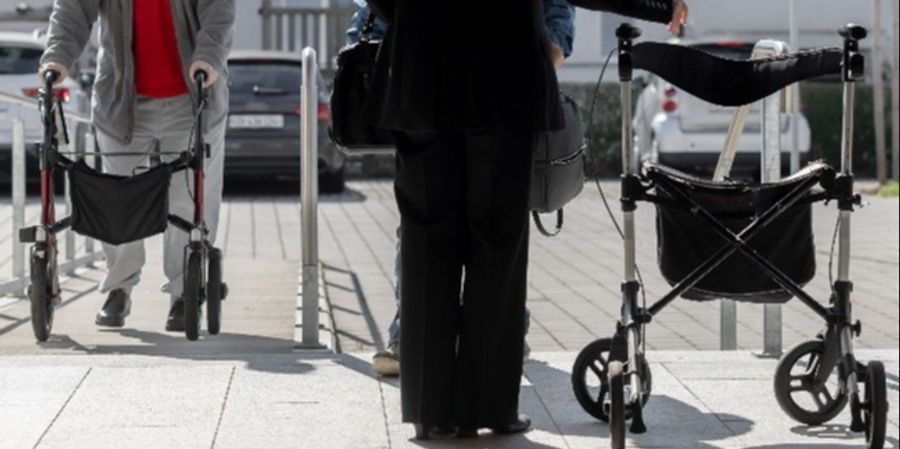 Auf einer Strasse sieht man zwei ältere Personen mit einem Rollatore.
