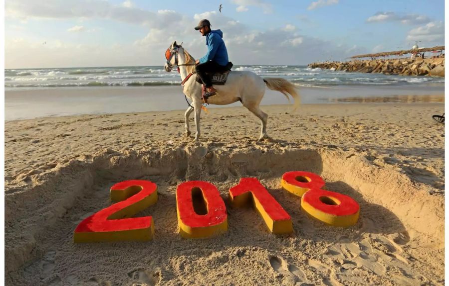 Ein Mann reitet am Strand in Gaza (Palästinensische Autonomiegebiete) hinter eine in Sand geformte Jahreszahl «2018» entlang.