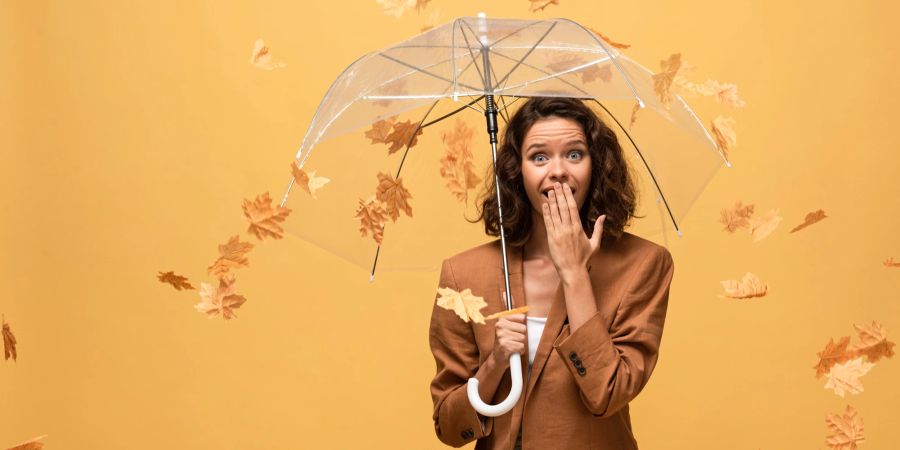 frau hält hand vor den mund, erschrocken, transparenter regenschirm, herbst-blätter fallen, gelber hintergrund