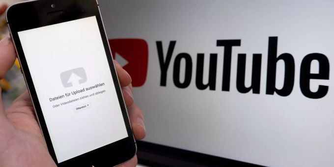 Ein Upload-Fenster der Video-Plattform Youtube ist auf einem Smartphone vor einem YouTube-Logo auf einem Bildschirm zu sehen. Artikel 13 Urheberrecht