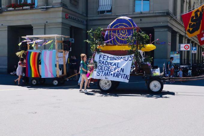 Zwei Wagen für den Frauenstreik fahren über den Bundesplatz. Der hintere mit Pride- und Trans-Flagge, der vordere mit einer Schnecke, eines der Symbole für den Streik. Schnecke Frauenstreik