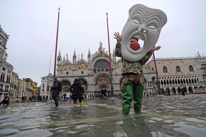Ein Mann steht in hohen Gummistiefeln und mit einer riesigen venezianischen Maske auf dem überfluteten Markusplatz. Hochwasseralarm in Venedig