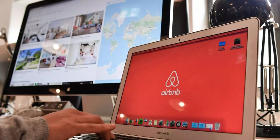 Auf einem Laptop und einem Bildschirm im Büro der Airbnb sind das Logo des Unternehmens und eine Auswahl von Unterkünften zu sehen.