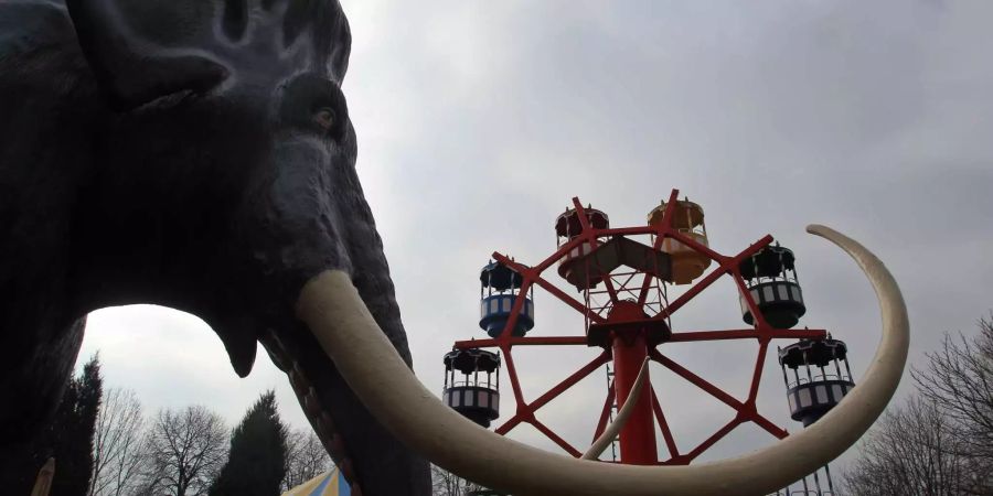 Ein künstliches Mammut und ein Kinderriesenrad stehen in einem Freizeitpark vor bedecktem Himmel.