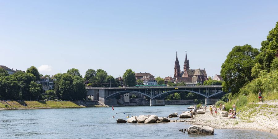 Die Wettsteinbrücke und das Münster in Basel. Am Rhein vergnügen sich Sonnenhungrige, beliebt ist auch das Schwimmen im Rhein.