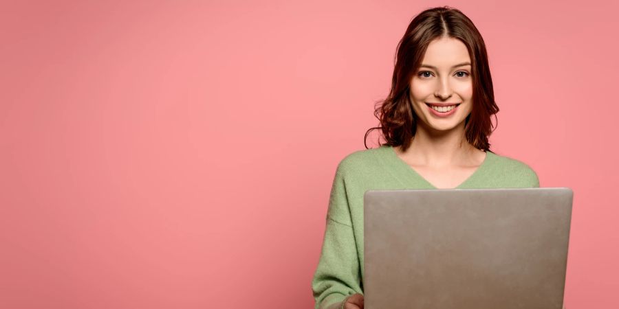 Eine Frau schaut lächelnd in die Kamera und hält dabei einen aufgeklappten Laptop.