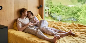 Mann und Frau im Bett vor grossen Fenstern mit Natur Blick.