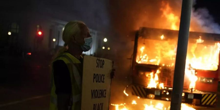 Ein Demonstrant steht mit einem Transparentvor einem brennenden Müllwagen. Foto: David Goldman/AP/dpa