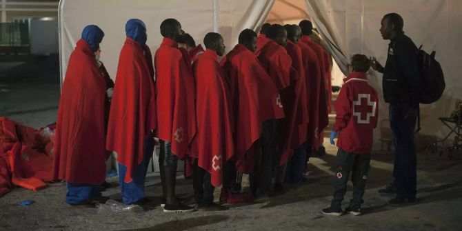 In rote Decken gehüllte Migranten stehen nach ihrer Ankunft im Hafen von Malaga in einer Schlange vor einem Zelt des Roten Kreuzes.