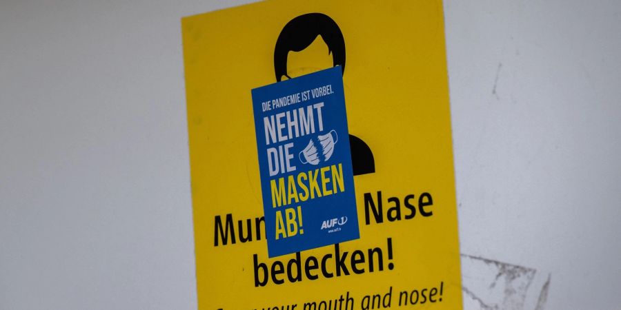 Ein Aufkleber mit der Aufschrift «Nehmt die Masken ab!» klebt über einem Schild, das zum Tragen einer Mund-Nasen-Maske auffordert. Die Mehrheit der Deutschen ist einer neuen Umfrage zufolge weiter für eine Isolationspflicht für Corona-Infizierte.