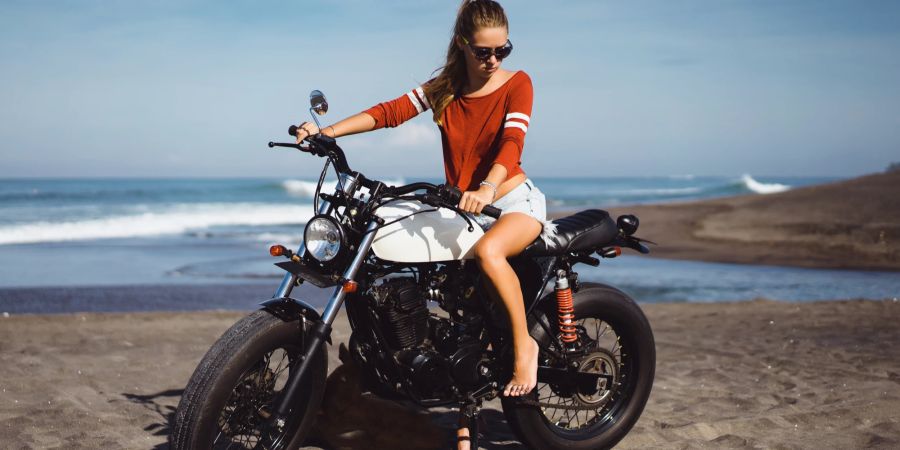 Frau in Shorts und Shirt ohne Helm auf einem Motorrad am Strand.