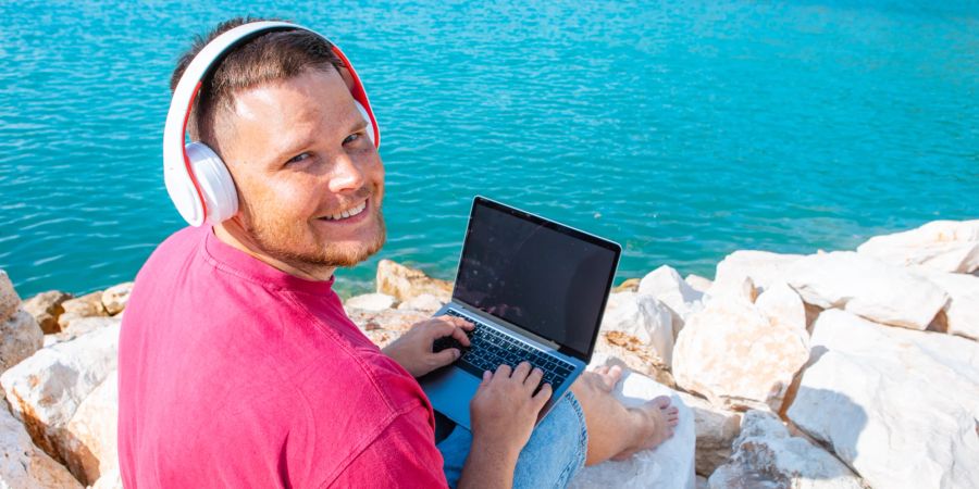 Mann mit Laptop und Kopfhörern am Meer.