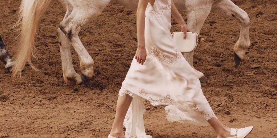 Model mit weissem KLeid im Sand mit Pferd.