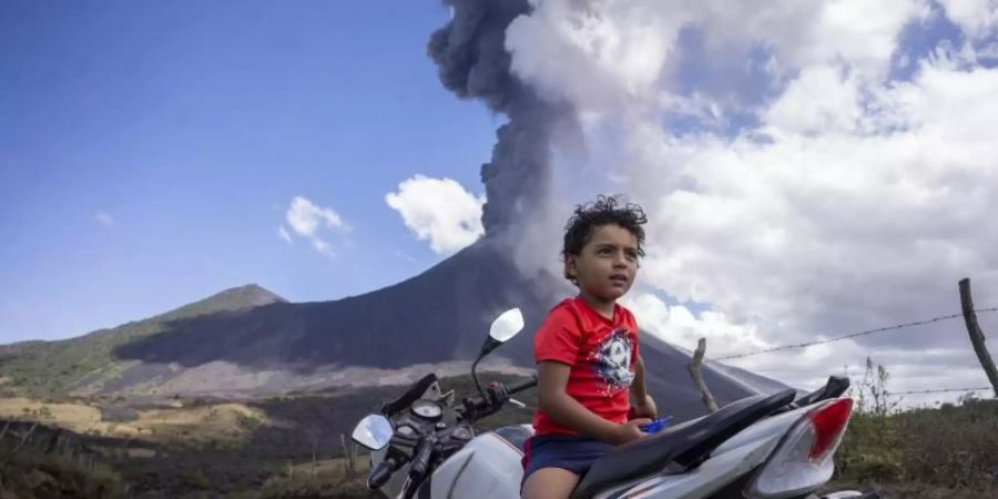 Ein Kind sitzt auf einem Motorrad, während im Hintergrund der Vulkan Pacaya ausbricht. Foto: Santiago Billy/AP/dpa