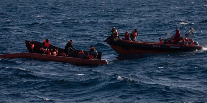 Mehrere Migranten sitzen in einem Boot im Mittelmeer während Rettungskräfte versuchen ihnen zu helfen.