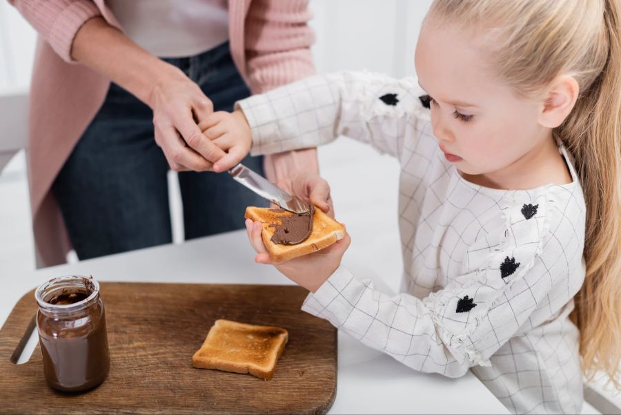 Kleines Mädchen verteilt Schokoladenpaste auf Brot.