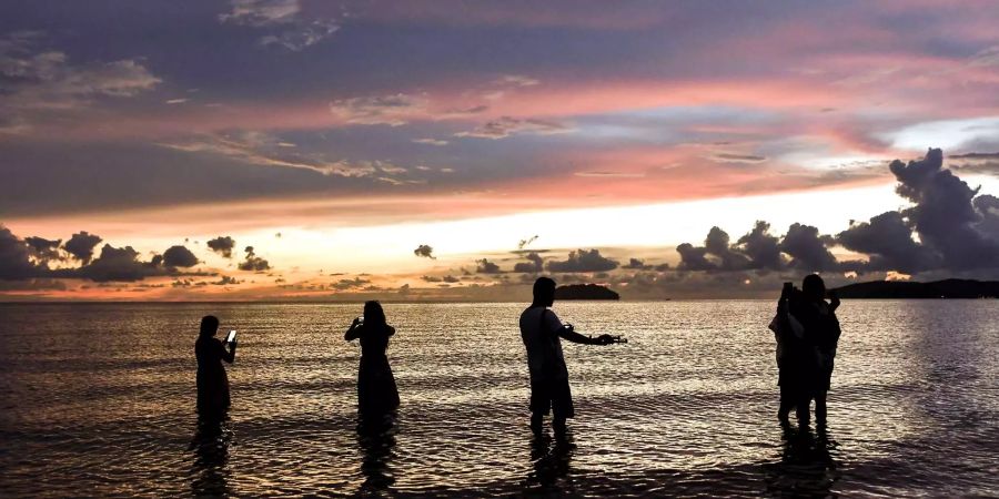 Touristen stehen am beliebten Strand von Tanjung Aru in Malaysien im Wasser und fotografieren den Sonnenuntergang.