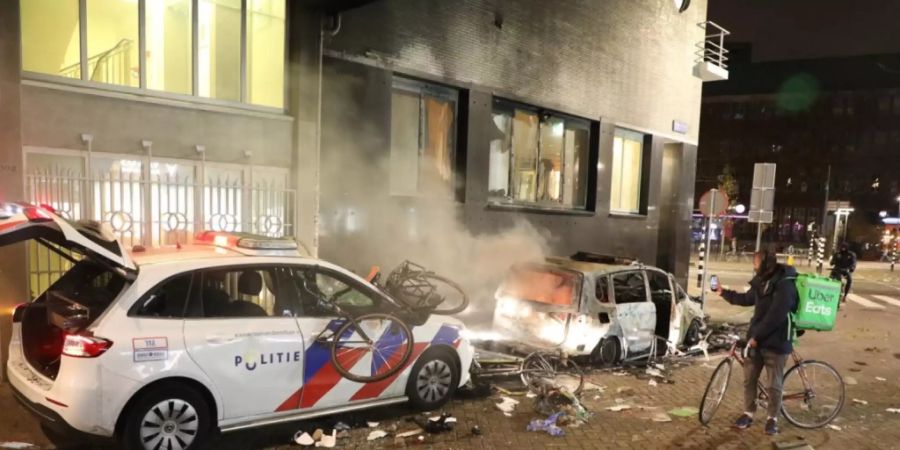 Ein Fahrradkurier macht ein Bild von einem ausgebrannten Auto. Daneben steht ein beschädigter Polizeiwagen. Foto: -/ANP/dpa