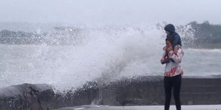 Eine Welle tritt über das Ufer während eine Frau mit einem Kind auf den Schultern an der Strandpromenade geht. Sturmtief «Ellen» trifft auf die Küste Irlands. Foto: Niall Carson/PA Wire/dpa