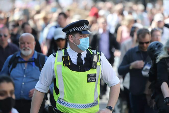 Ein Polizist mit Mund-Nasen-Schutz steht inmitten einer Gruppe von Demonstranten. Coronavirus London