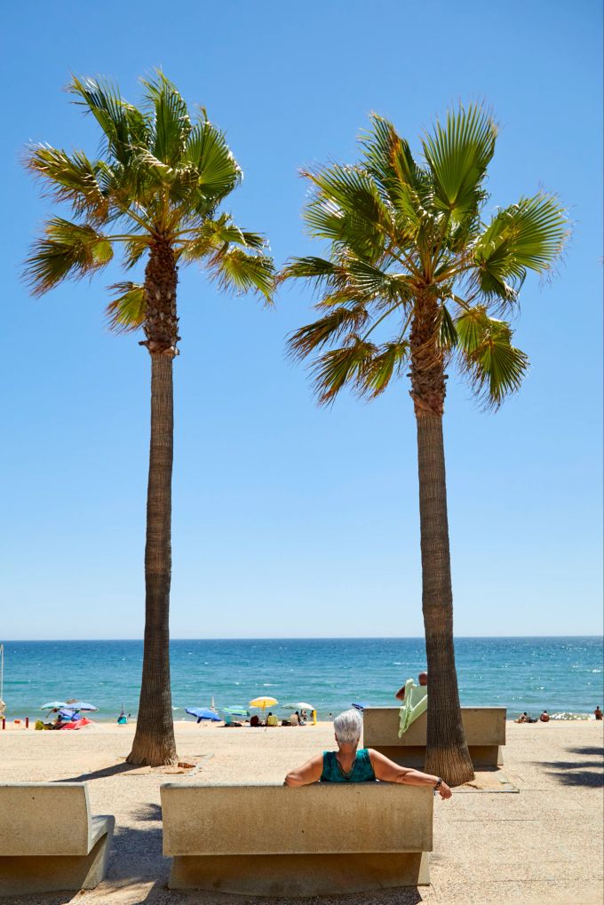 Bank Mann Zement vor Strand mit Palmen Meer blauer Himmel