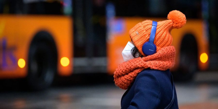 Eine Frau sitzt mit FFP2-Maske, dicker Mütze und Kopfhörern an einem Busbahnhof in Hamburg.