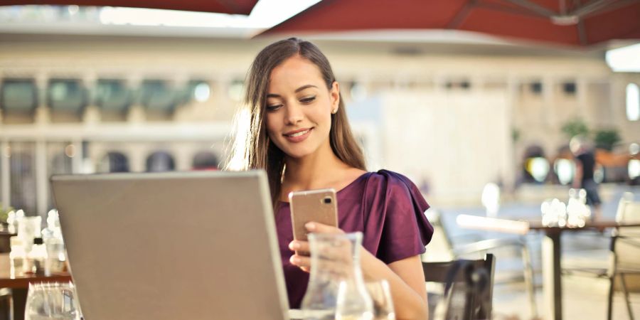 Eine Frau sitzt an einem Tisch vor ihrem Laptop in einer Küche und schaut auf ein Smartphone.