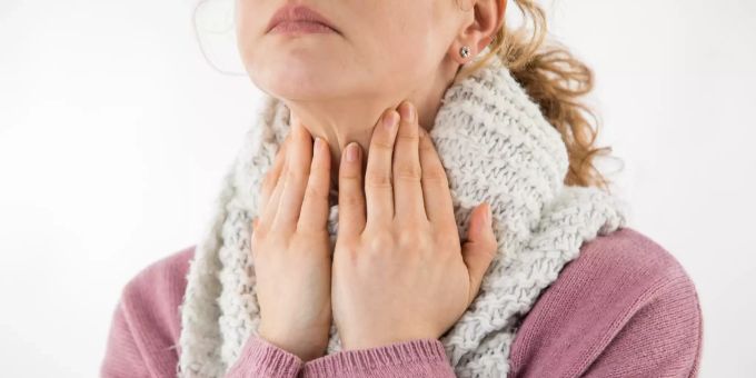 Eine heisere Stimme kann verschiedene Ursachen haben. Manchmal stecken kleine Knötchen auf den Stimmlippen dahinter. Frau Hals Hände Schal weiss Pullover alrosa