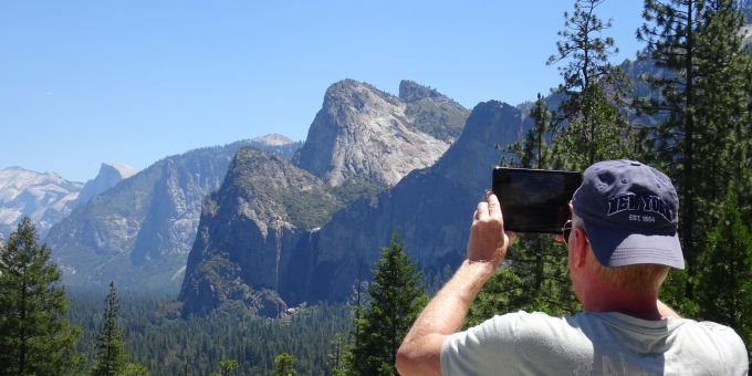 In den Yosemite-Nationalpark kann man im Sommer 2022 nicht ohne Weiteres mit dem Mietwagen hineinfahren: Tagesausflügler müssen im Internet für ihr Auto reservieren. Mann Smartphone Gebirge Wald Nadelbäume Sonne