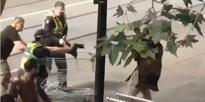 Das Standbild aus einem Video zeigt Michael Rogers (l, mit Einkaufswagen) und Polizisten, die einen Mann (r), der mit einem Messer bewaffnet ist, überwältigen zu versuchen. Das Standbild aus einem Video zeigt Michael Rogers (l, mit Einkaufswagen) und Polizisten, die einen Mann (r), der mit einem Messer bewaffnet ist, überwältigen zu versuchen.