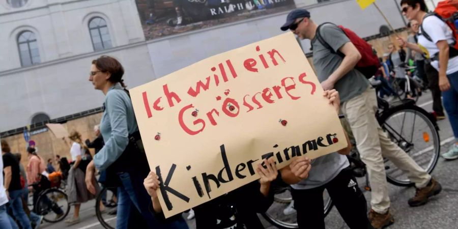 Teilnehmer einer Demonstration für bezahlbaren Wohnraum und gegen soziale Ausgrenzung unter dem Motto #ausspekuliert ziehen mit einem Plakat mit der Aufschrift «Ich will ein grösseres Kinderzimmer» durch die Ludwigsstrasse und gehen an einem grossen Werbeplakat an einer Hauswand vorbei.