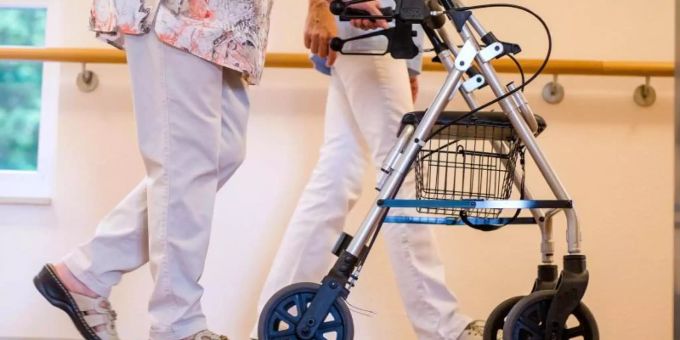 Eine Pflegekraft geht in einem Pflegeheim mit einer älteren Dame über einen Korridor. Pflege