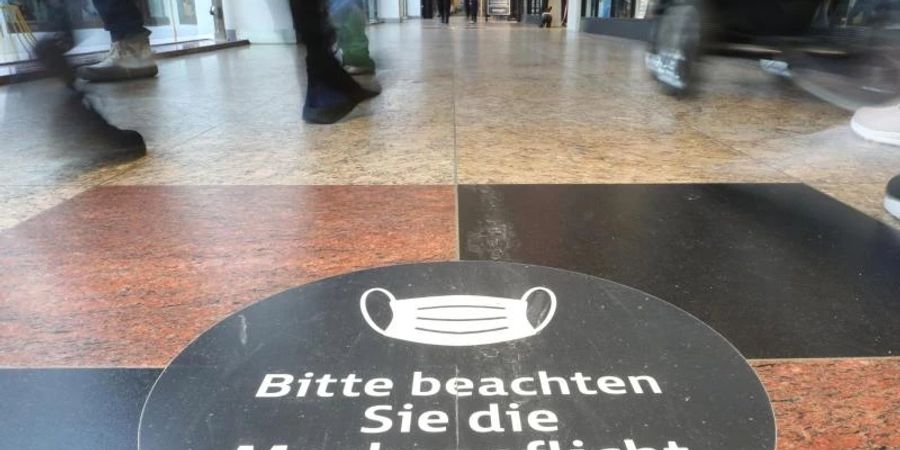 Passanten laufen durch ein Einkaufszentrum auf dessen Fussboden ein Hinweisschild zur Maskenpflicht aufgeklebt ist. Foto: Danny Gohlke/dpa
