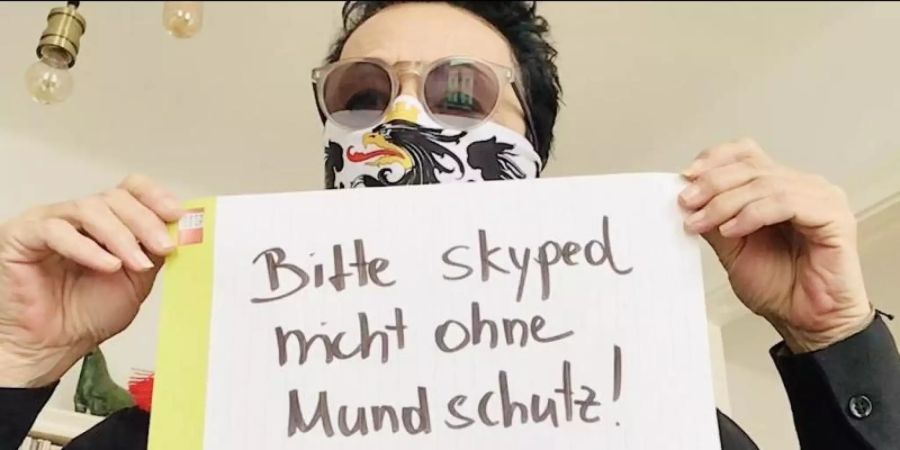 Ein Mann mit Mundschutz hält in einem Ausschnitt aus der "Corona-Rolle" ein Schild mit der Aufschrift "Bitte skyped nicht ohne Mundschutz" hoch. Foto: --/EchtzeitExperiment/dpa