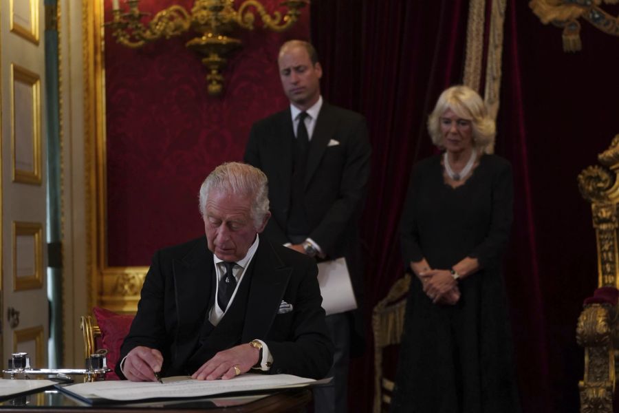 King Charles unterschreibt die Proklamation. Camilla und Prinz William im Hintergrund.