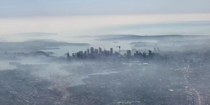 Auf dem mit einem Smartphone durch ein Flugzeugfenster aufgenommenen Foto liegen Rauchschleier über der Skyline von Sydney. Buschbrände in Australien