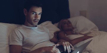 Mann mit Laptop und schlafender Freundin schaut verdächtig in Kamera