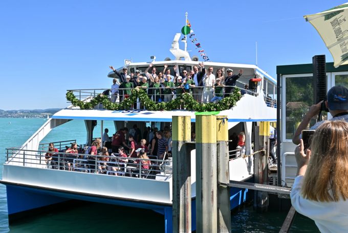Mehrere Festgäste winken zum Abschluss der Taufe des elektrobetriebenen Katamarans auf den Namen «Insel Mainau» am Landesteg vor der Insel Mainau im Bodensee vom Oberdeck. Foto: Felix Kästle Neuer E-Katamaran Insel Mainau