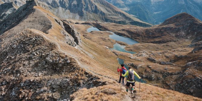 Zwei Männer auf einem Panoramaweg in den Alpen mit Seen im Hintergrund an einem sonnigen Herbsttag.