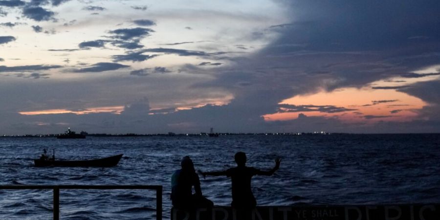 ARCHIV - Menschen sitzen auf der Uferpromenade, während die Sonne in Georgetown, Guyana, untergeht. Knapp eine Woche nach dem schweren Fährunglück vor der Küste Guyanas sind inzwisc...