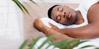 schwarzer mann schläft in weisser bettwäsche, pflanze im vordergrund