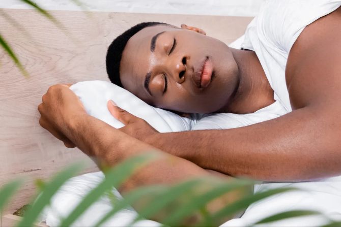 schwarzer mann schläft in weisser bettwäsche, pflanze im vordergrund