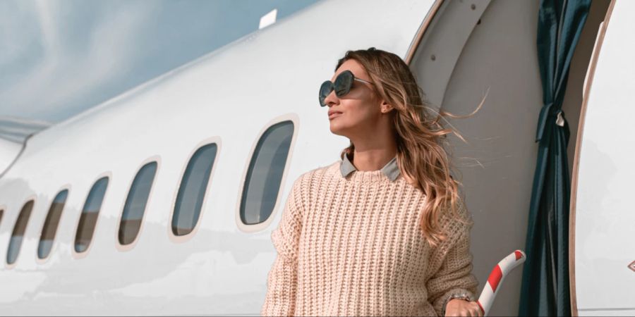 Frau mit Sonnenbrille und rosa Strickpullover stht auf der Gate-Treppe vor einem Flugzeug.