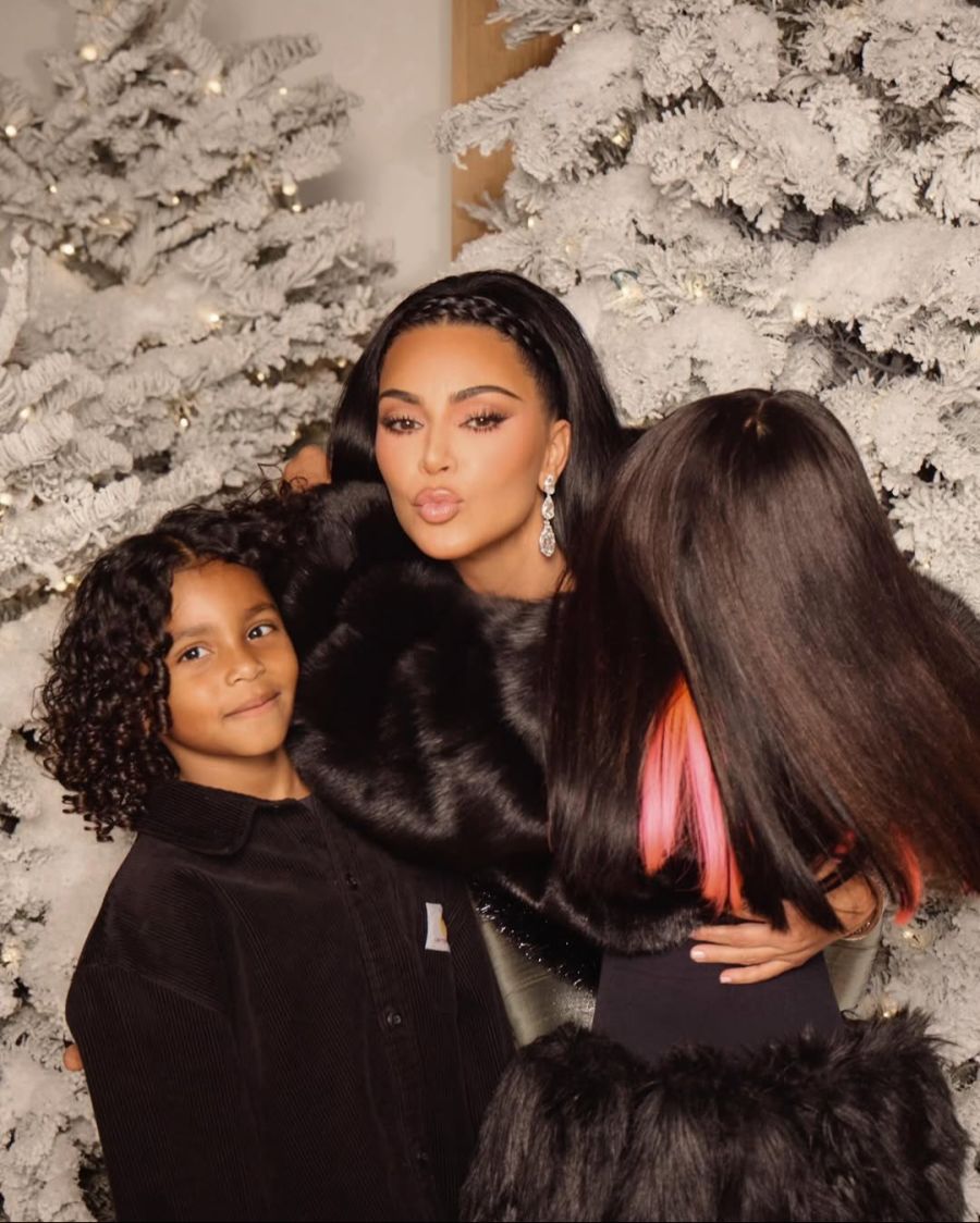 Kim Kardashian mit Tochter Chicago und Sohn Saint vor schneebedeckten Christbäumen. Alle posieren im Partnerlook in Schwarz.