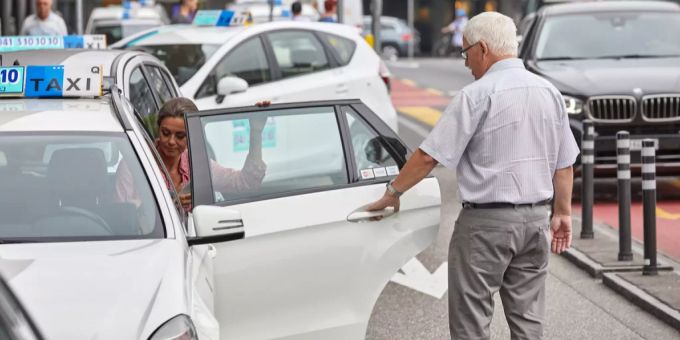 Ein Taxichauffeur im Rentenalter hilft einer Frau ins Fahrzeug. (Symbolbild) taxi rentenalter
