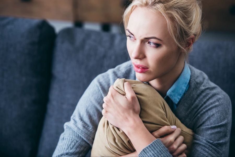 Ängstliche und traurige Frau sitzt auf Couch und hält Kissen fest.