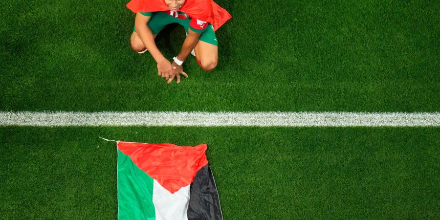 Marokkos Abdelhamid Sabiri feiert neben einer palästinensischen Flagge auf dem Rasen hockend den Sieg seiner Mannschaft.