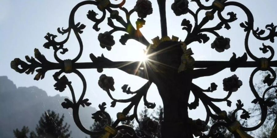 Die Sonne leuchtet durch ein Metallkreuz auf einem Friedhof. Foto: Angelika Warmuth/dpa