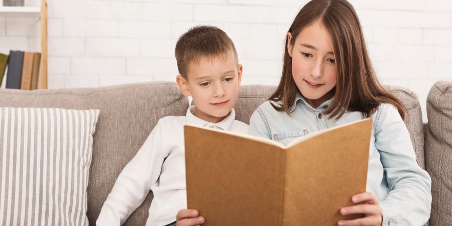 Schwester liest kleinem Bruder ein Buch vor.