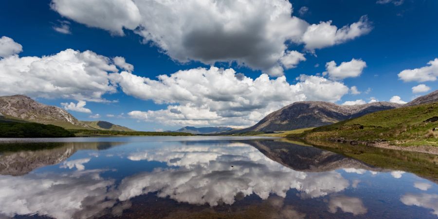 Blick auf einen See mit Spiegelung von Wolken und Himmel. Umliegende grüne Hügel Irlands.